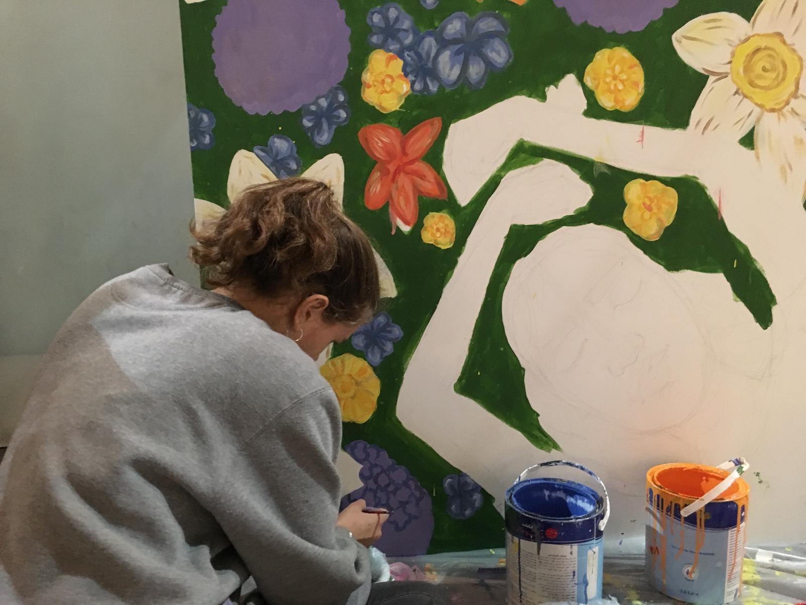 Phoebe sits on the ground, bent over a section of a mural she created at the mall. It depicts a woman laying in a field of flowers, her arms stretched above her head.