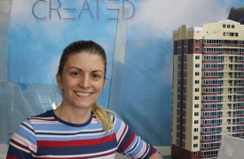 Irina, smiling in the center, stands next to a model building with her company's sign, 3D Create, in the background