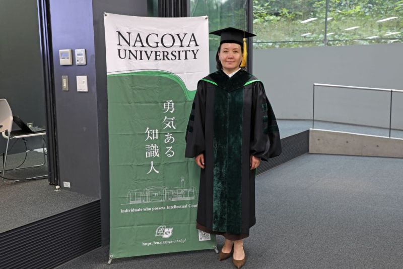 Woman poses for photo in graduation cap and gown in front of a banner that reads "Nagoya University"