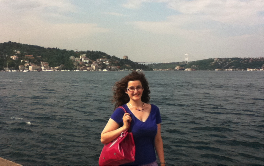 August poses in Istanbul with the wind blowing her hair as she holds a red handbag, standing on the short of the Bosphorus Sea.