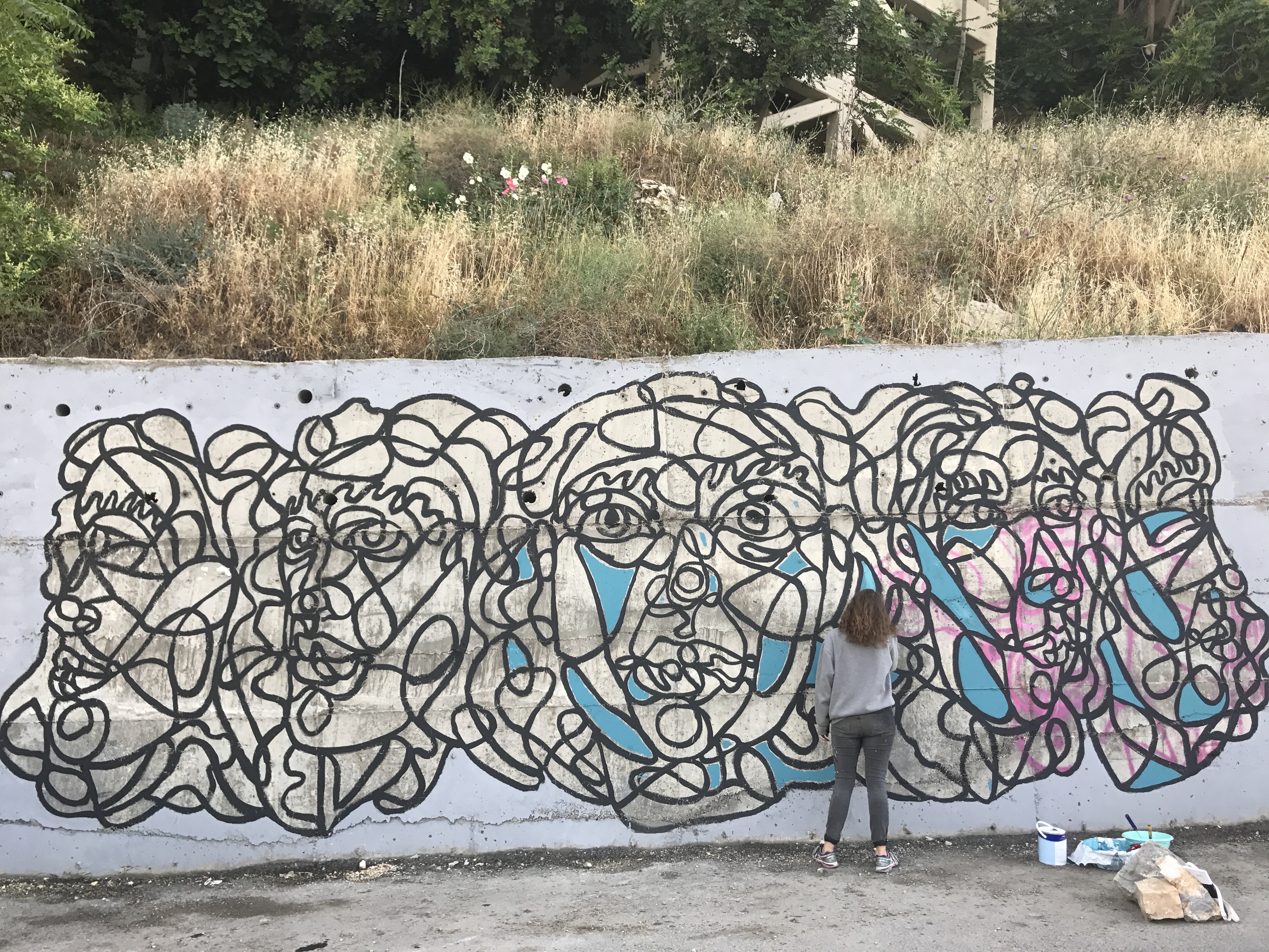 Phoebe Rossman stands in front of a concrete wall, painting a section of her mural. Abstract faces of women, with intersecting lines, have been drawn and she is slowly adding color blocks to the piece.