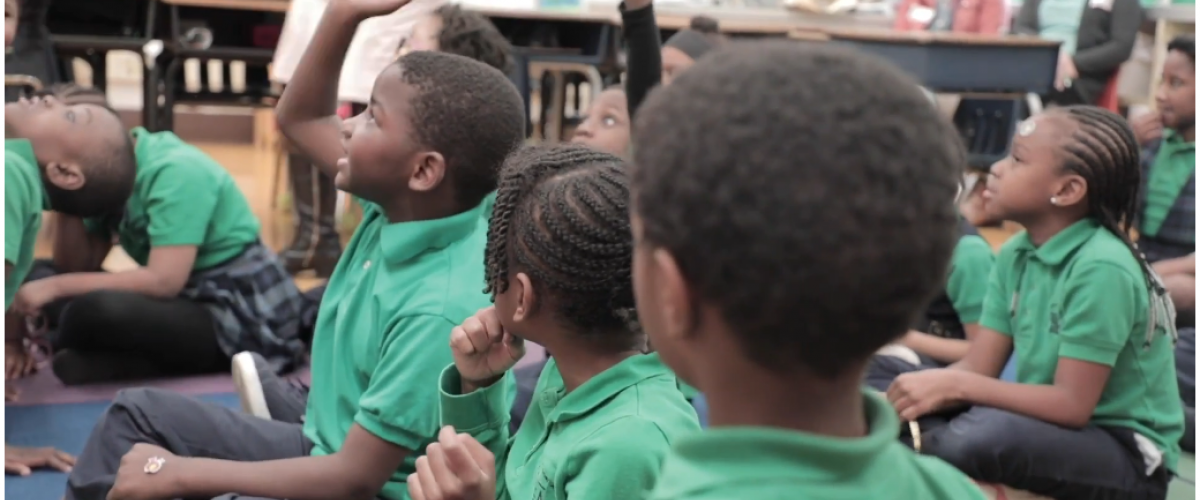 Young students in green sit on the classroom floor