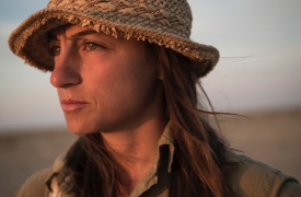 Anush, wearing a straw hat and an olive, long sleeve button down shirt, looks off into the sunset.