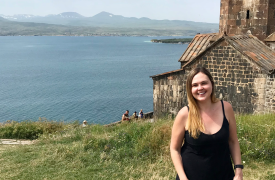 Jeni, wearing a black sundress, poses at Lake Sevan, with the lake and a stone building in the background.