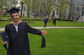 Egzon, dressed in a graduation cap and gown, poses on the lawn of his university.