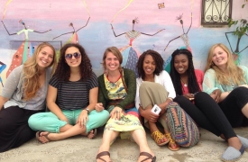 A group of young women sit against a colorful wall, smiling and relaxing