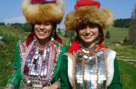 Two women wearing traditional dress, including fur hats, pose together on a hillside