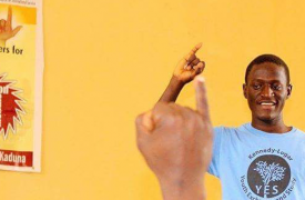 Against a sunny yellow wall, Nehemiah from Nigeria teaches sign language to fellow deaf students