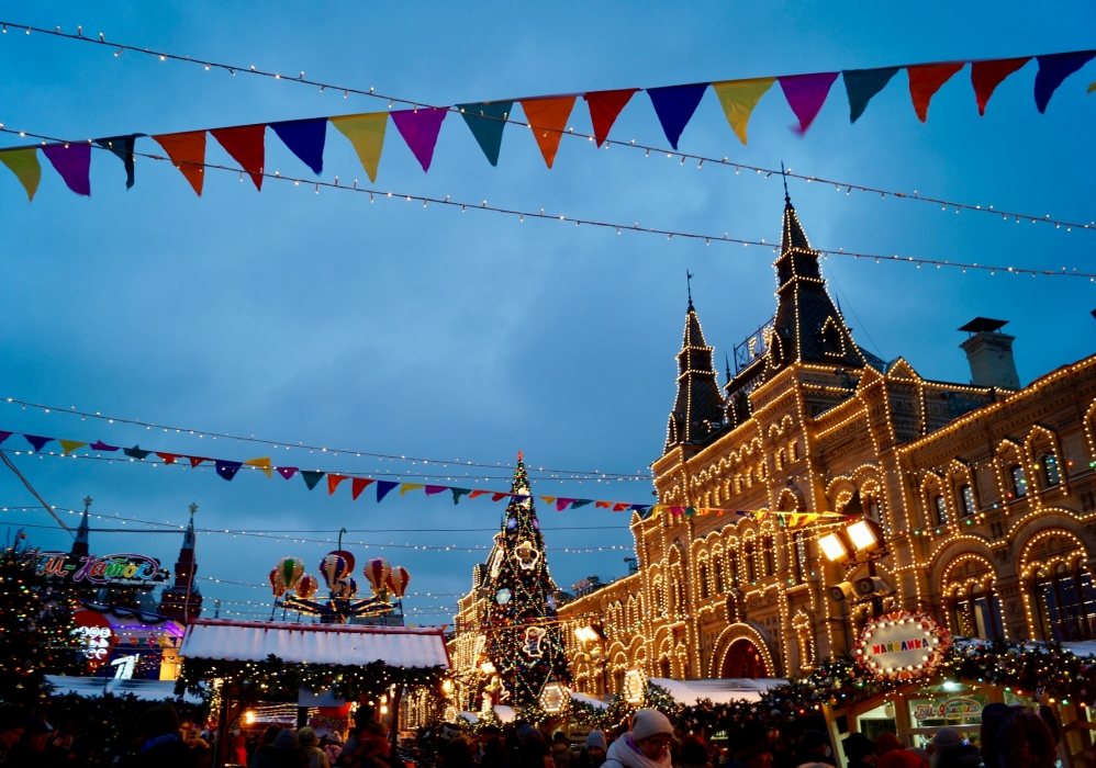 A large, stone building lit up with white, string lights, is the back drop to a holiday market in a Russian city