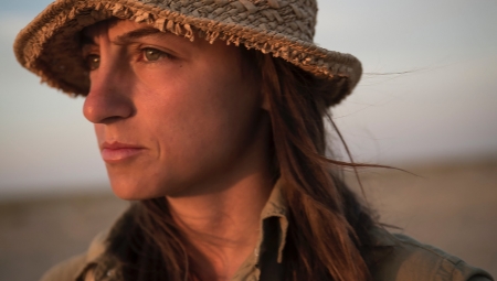 Anush, wearing a straw hat and an olive, long sleeve button down shirt, looks off into the sunset.