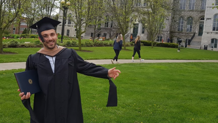 Egzon, dressed in a graduation cap and gown, poses on the lawn of his university.