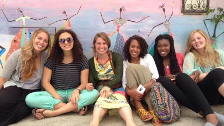 A group of young women sit against a colorful wall, smiling and relaxing