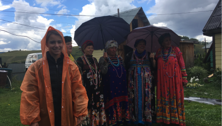 Isabella, in an orange rain jacket, smiles in front of a Kazakstani choir dressed in traditional clothing