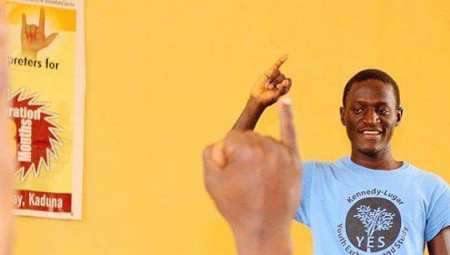 Against a sunny yellow wall, Nehemiah from Nigeria teaches sign language to fellow deaf students